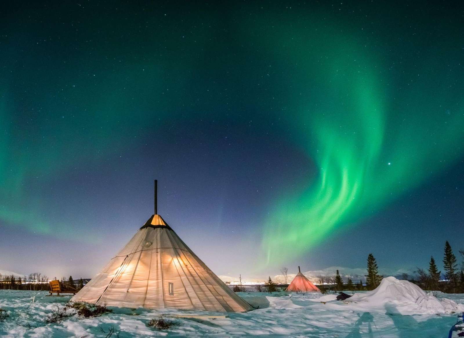 Aurora over lit Sámi tents in the snow at Kilpisjärvi, northwest Finnish Lapland